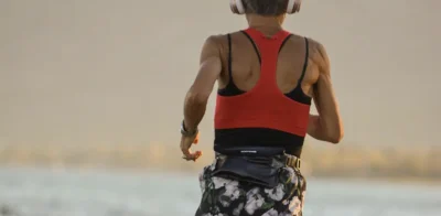 Een fitte vrouw die bij zonsondergang langs de kust rent, met een koptelefoon op, een rood sporttopje en een korte broek met een patroon.