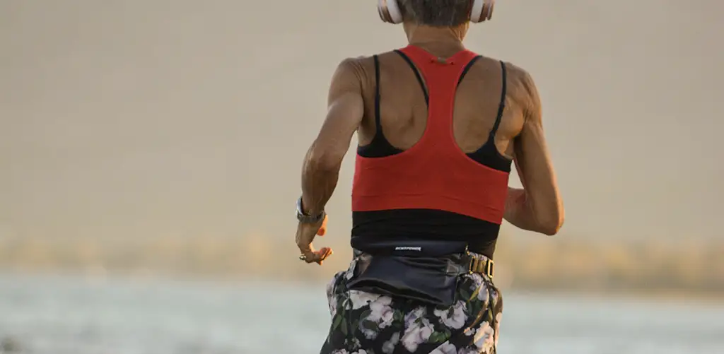 Een fitte vrouw die bij zonsondergang langs de kust rent, met een koptelefoon op, een rood sporttopje en een korte broek met een patroon.