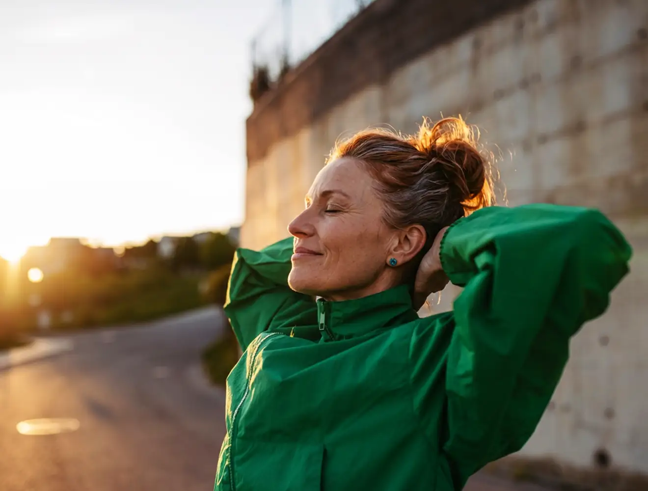 Eine reife Frau in einer grünen Jacke, die bei einem sonnigen Training im Freien mit geschlossenen Augen Dehnübungen macht – ganz entspannt und voller Energie