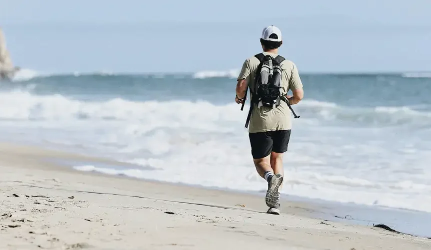 A man runs alone along a quiet beach beside the ocean, wearing a backpack as waves wash ashore under a pale blue sky.