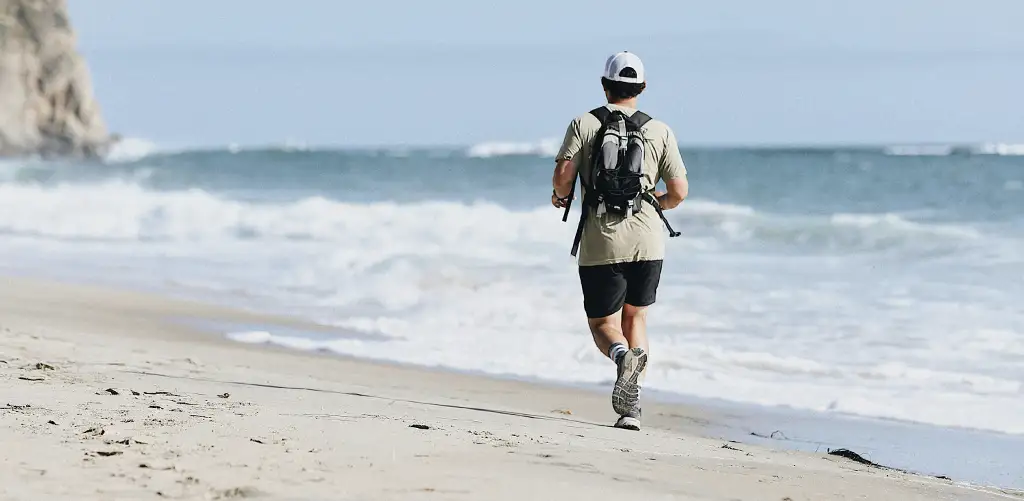 A man runs alone along a quiet beach beside the ocean, wearing a backpack as waves wash ashore under a pale blue sky.