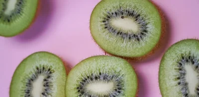 Close-up of several sliced green kiwis arranged on a pink background, showing their bright green flesh, white centers, and small black seeds in a radial pattern.