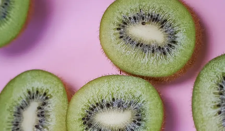 Primer plano de varios kiwis verdes cortados en rodajas y dispuestos sobre un fondo rosa, en el que se aprecia su pulpa de color verde brillante, el centro blanco y las pequeñas semillas negras dispuestas en forma radial.
