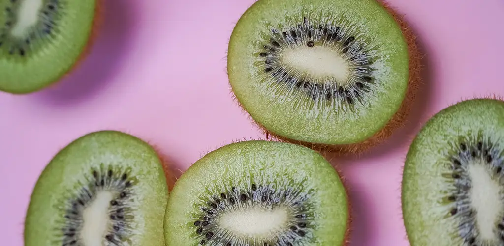 Nahaufnahme mehrerer aufgeschnittener grüner Kiwis, die auf einem rosa Hintergrund angeordnet sind und deren leuchtend grünes Fruchtfleisch, weiße Kerne und kleine black in einer radialen Anordnung zu sehen sind.