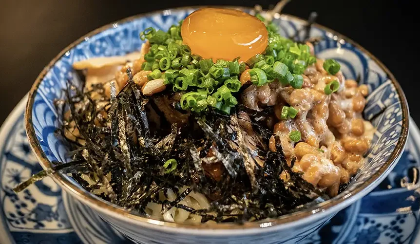 Un cuenco de natto con yema de huevo cruda, cebollas verdes picadas y algas desmenuzadas por encima, servido sobre arroz.