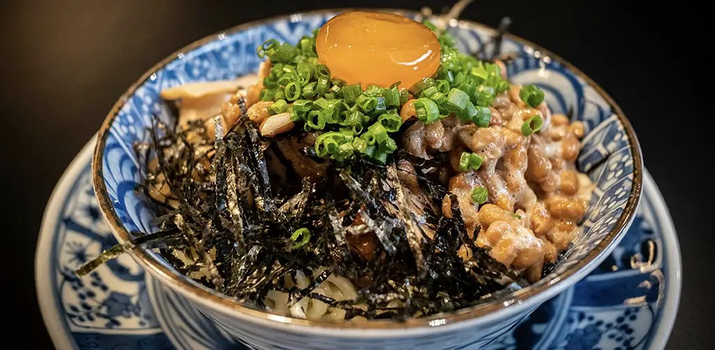A bowl of natto topped with raw egg yolk, chopped green onions, and shredded seaweed, served over rice.