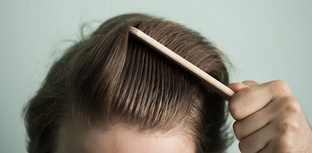 Close-up of a person combing back their light brown hair with a wooden comb, revealing the hairline and scalp.