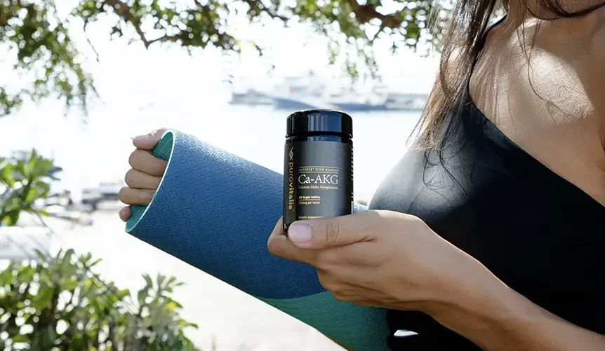 Woman holding a bottle of calcium alpha-ketoglutarate (Ca-AKG) supplement outdoors with a yoga mat