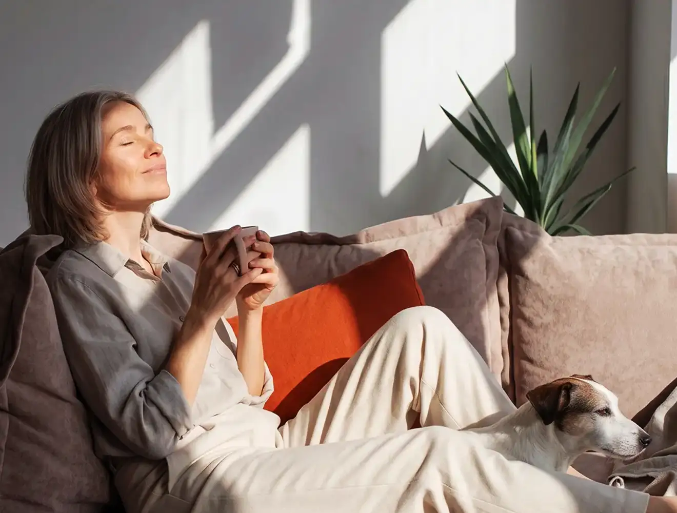 Woman relaxing on a sofa in natural sunlight while holding a cup of coffee, with a dog resting beside her in a calm and cozy home environment.