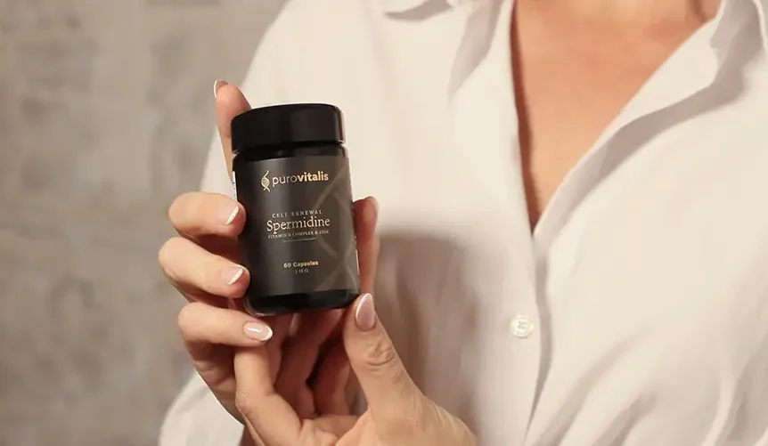 En person, der holder en flaske Purovitalis Spermidine-kosttilskud foran en neutral baggrund, hvilket fremhæver den liposomale formel for longevity og den eksklusive emballage.