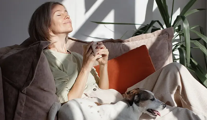 Kvinde, der slapper af i en sofa i naturligt sollys med en kop kaffe i hånden, mens en hund hviler ved siden af hende i et roligt og hyggeligt hjem.