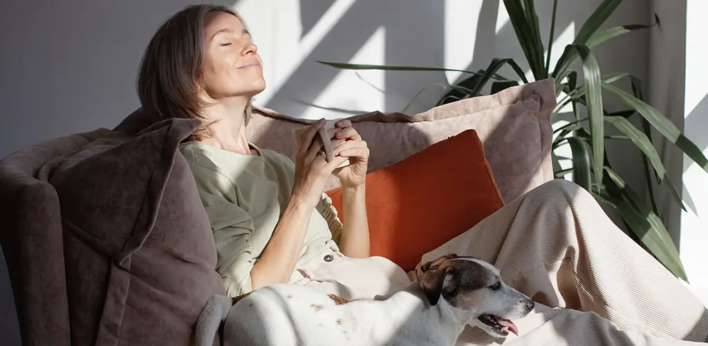 Woman relaxing on a sofa in natural sunlight while holding a cup of coffee, with a dog resting beside her in a calm and cozy home environment.
