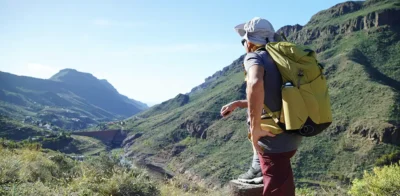 Man hiking in a mountainous landscape with a large backpack, looking out over a sunny valley during an outdoor trekking adventure.