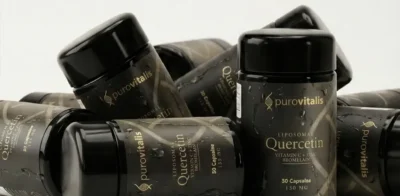 Varias botellas negras del suplemento «Purovitalis Liposomal Quercetin», con letras doradas que destacan los ingredientes añadidos —vitamina C, zinc y bromelina—, dispuestas sobre un fondo claro con gotitas de agua en el envase.
