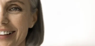 Primo piano del lato destro del viso di una donna matura sorridente, con le naturali rughe dell'età intorno agli occhi e alla bocca, su uno sfondo bianco brillante con ampio spazio vuoto sulla destra.