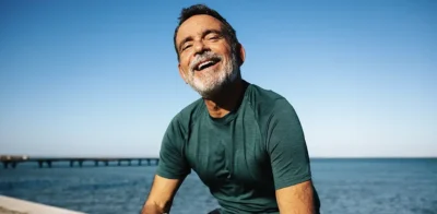 A smiling, athletic senior man, representing health and vitality after exercise, is sitting by the water under a clear blue sky.