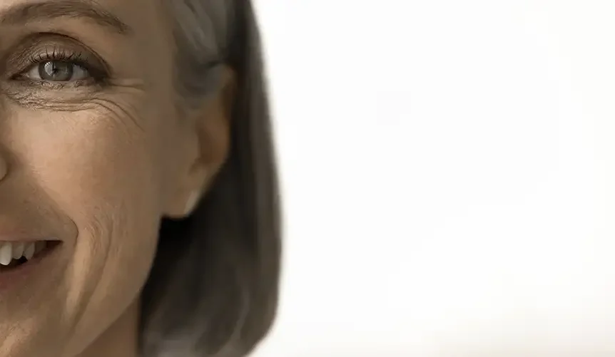 Close-up of the right side of a smiling mature woman's face with natural age lines around her eye and mouth, set against a bright white background with ample negative space on the right.