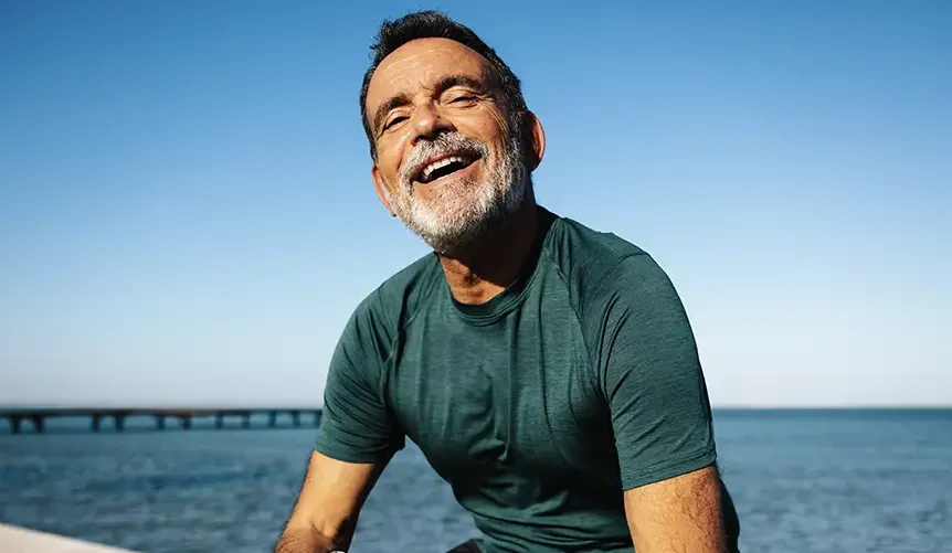 A smiling, athletic senior man, representing health and vitality after exercise, is sitting by the water under a clear blue sky.