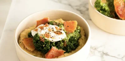 A healthy bowl of polenta topped with sauteed kale, blood orange slices, and a poached egg on a marble surface.