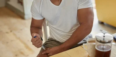 A muscular man administering a GLP-1 injection pen into his thigh while seated at a wooden table next to a coffee cup.
