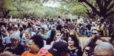 Un plano general de un grupo numeroso y variado de adultos sentados en sillas plegables blancas al aire libre, en un día soleado bajo unos árboles verdes.